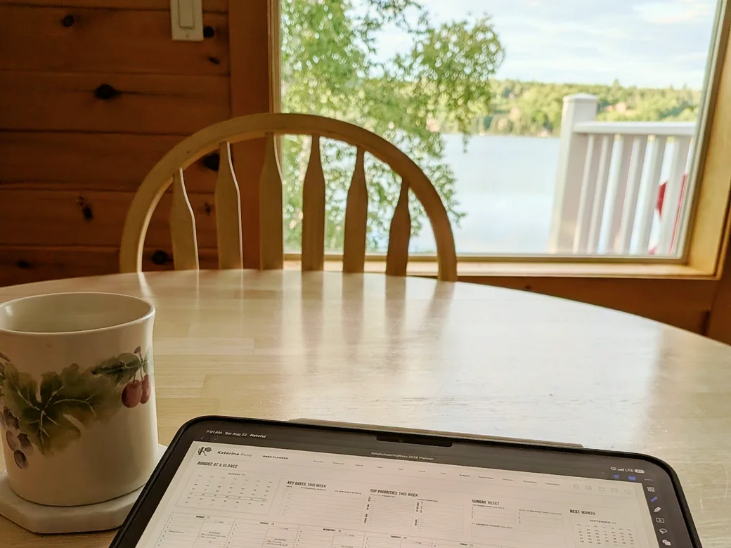 A tablet showing a weekly planner layout resting on a light wood table beside a painted ceramic mug of coffee, with a still lake and a wooden deck railing visible through the cabin window behind.