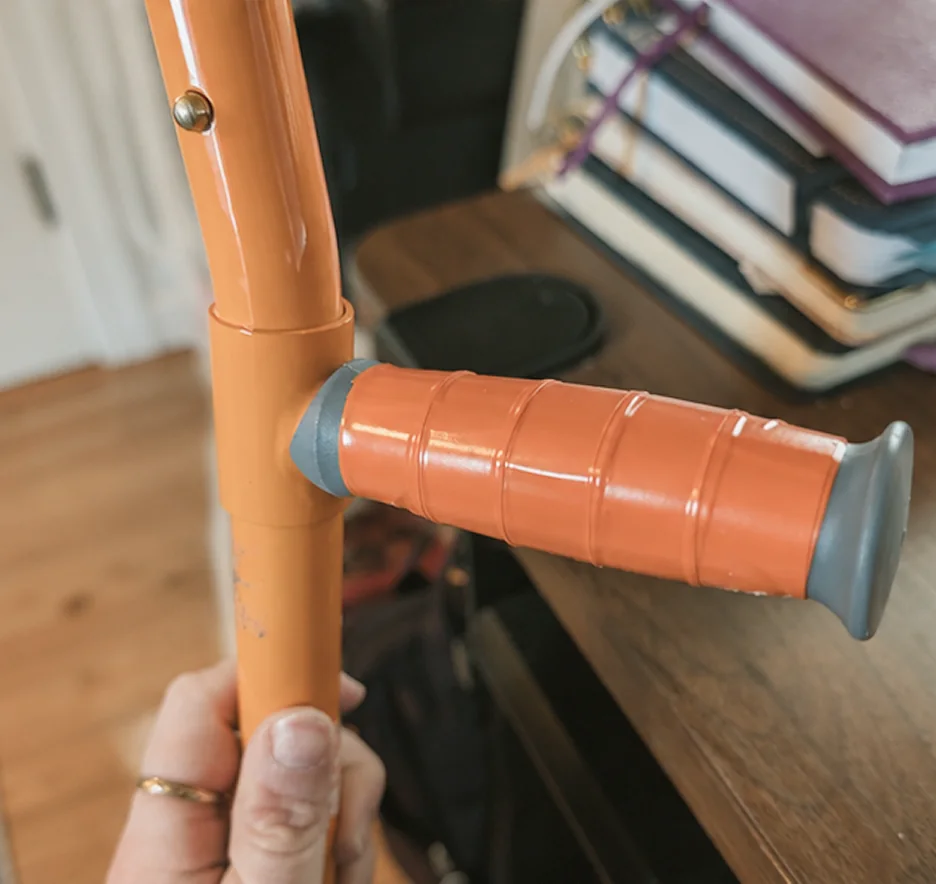 Close-up of an adaptive curling delivery stick held in one hand. A copper-orange grip capped in grey rubber is mounted crosswise near the top of the shaft, so the hand pushes across the stick rather than along it.
