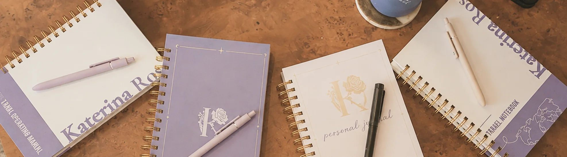 Four spiral-bound notebooks laid out on a wood table, each one designed differently: a lavender personal operating manual, a purple journal with a rose monogram, a cream personal journal, and a travel notebook printed with a world map. A pen rests on each.