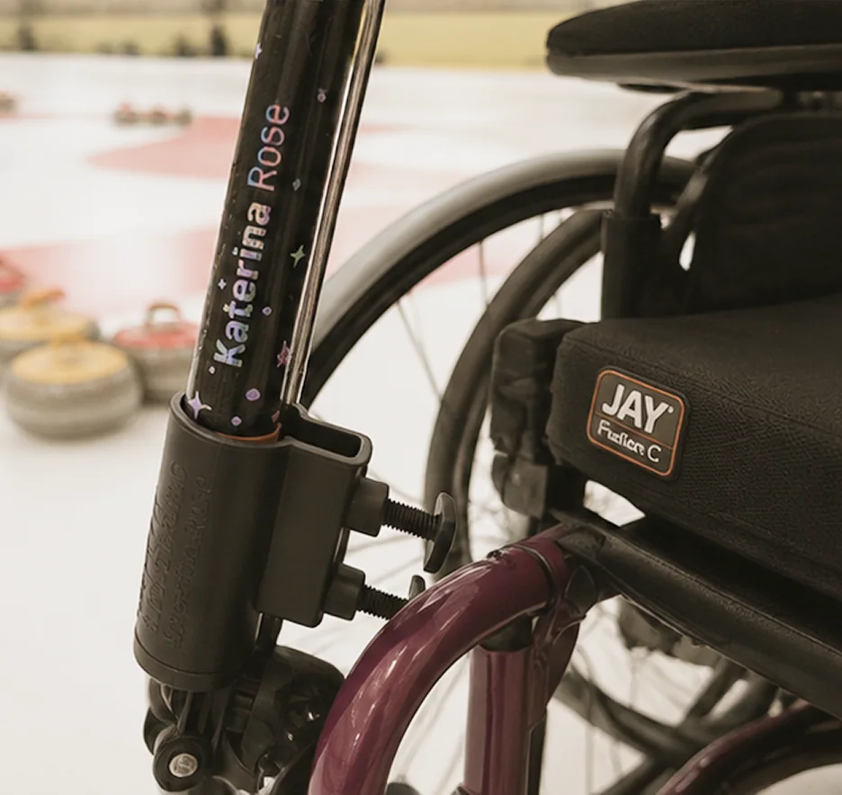 A curling delivery stick, personalized with the name Katerina Rose, clamped to the frame of a manual wheelchair by a black bracket. Behind it, curling stones rest on the ice near the painted rings of the house.